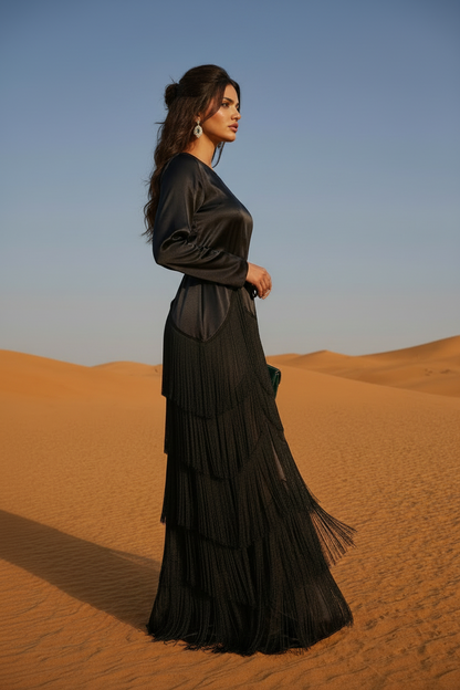 Woman in a black dress witb fringe standing in the desert with a clear blue sky.